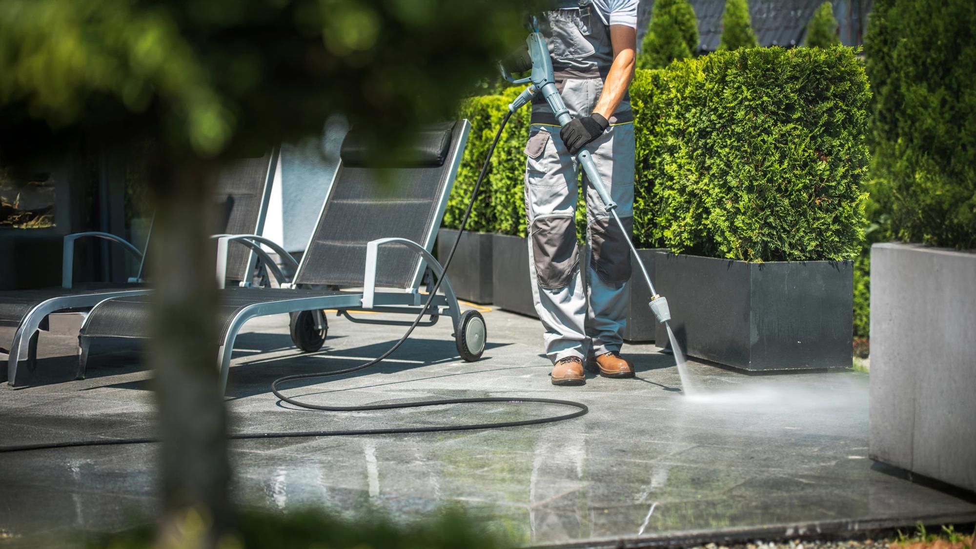 A person in gray overalls using a pressure washer on a stone patio, with lounge chairs and neatly trimmed hedges in the background.