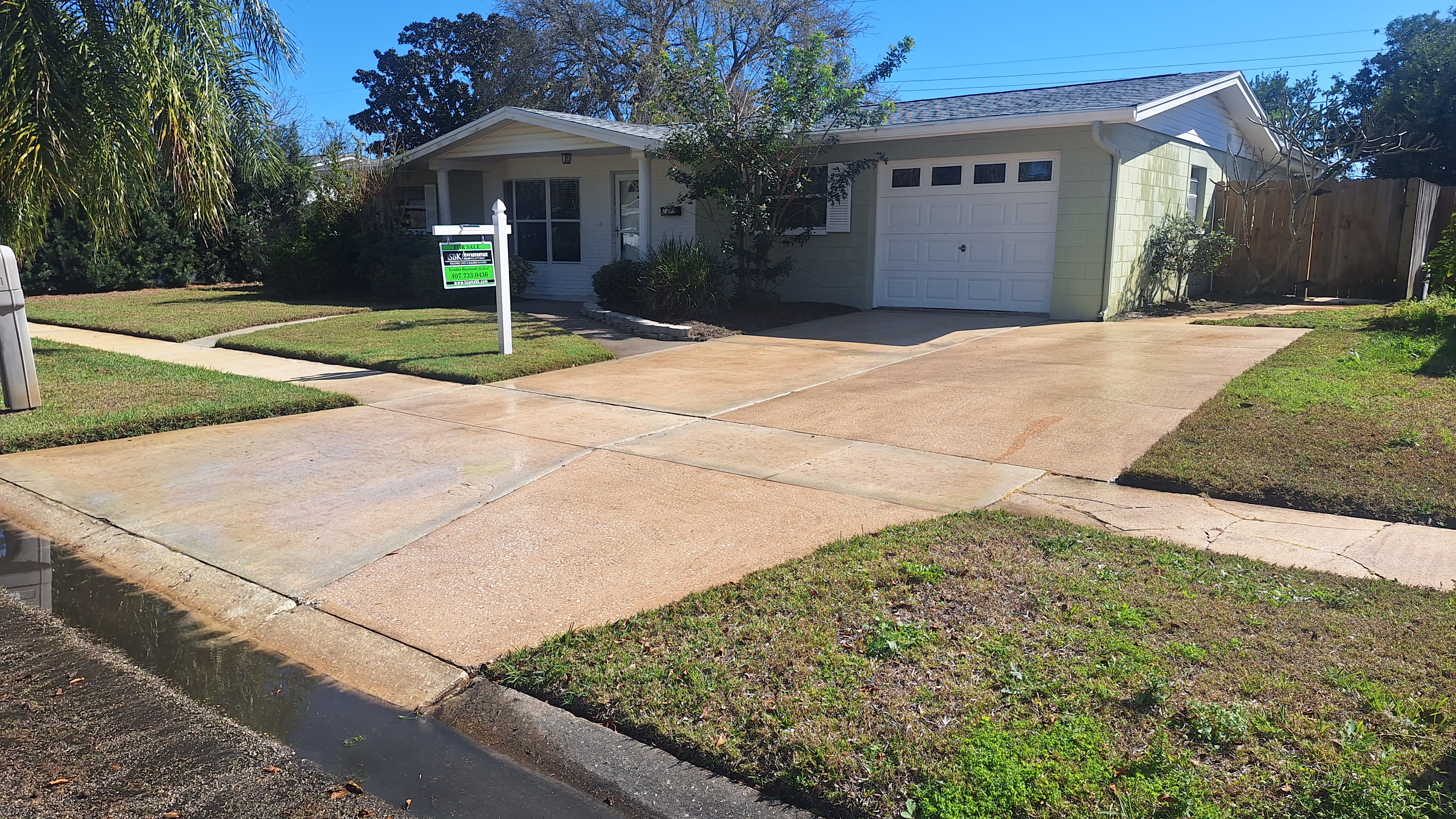 A person in a gray cap, blue overalls, and yellow rubber boots is using a pressure washer on a driveway near a modern garage. The pressure washer is on the ground beside them, with a hose connected. There are potted plants nearby and wet spots on the pavement.